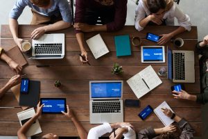 A group of people sit around a wooden table with laptops, tablets, notebooks, and mobiles, collaborating and discussing work; digital devices display blue screens with graphs and text.