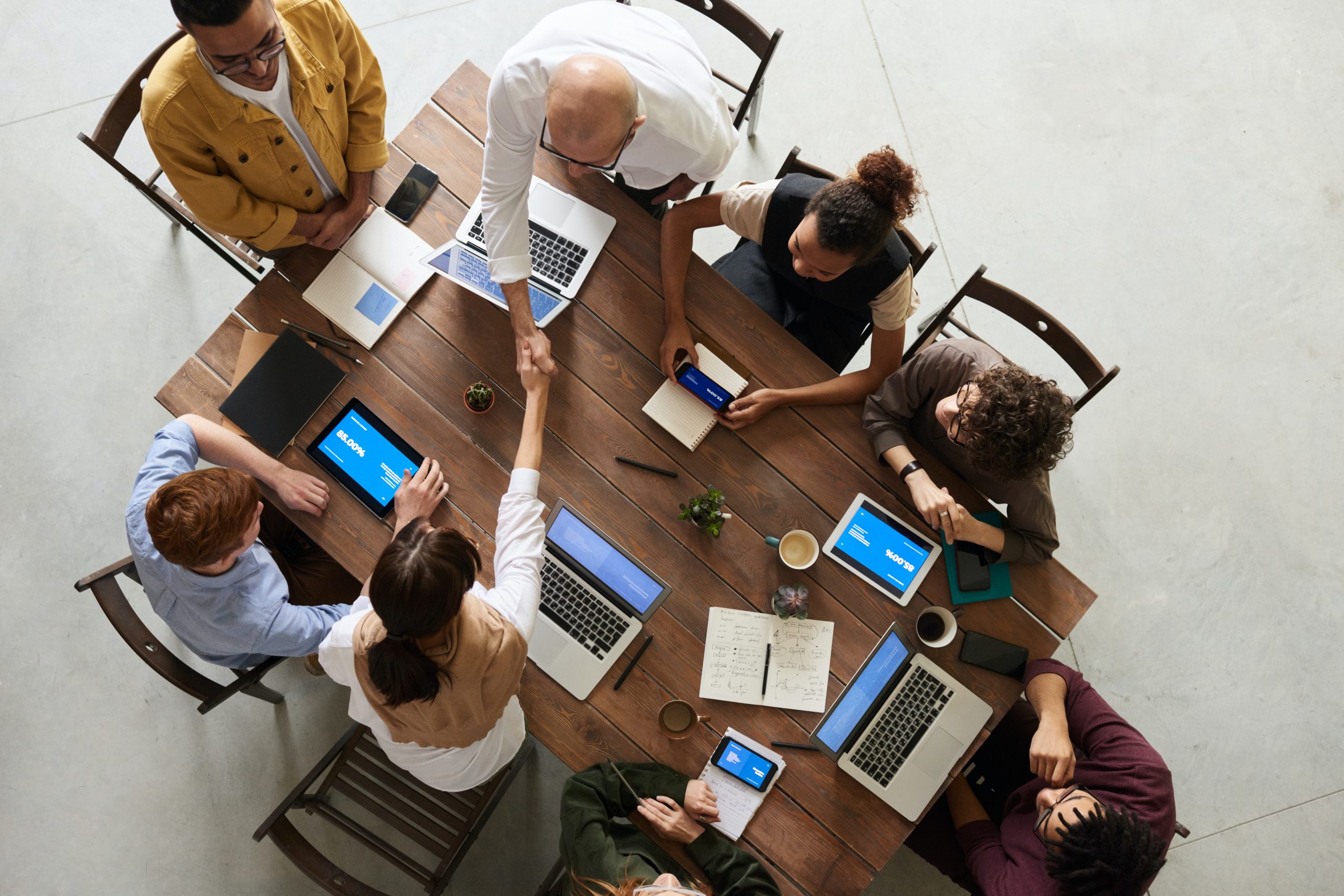 A group of people sit around a wooden table with laptops, notebooks, and coffee cups, while two people in the centre shake hands, viewed from above.